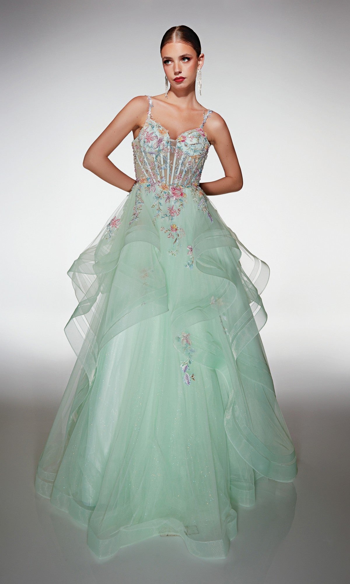 A woman poses gracefully in the Alyce 62201, a light green, floor-length formal dress featuring sheer layered tulle and delicate 3D floral appliques, set against a plain background.